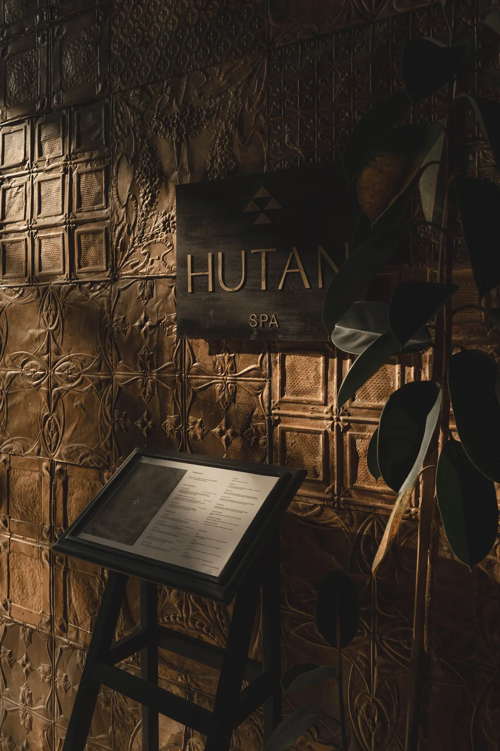 The brass Hutan Spa sign on a pressed-metal wall, beside a rubber plant and the menu stand