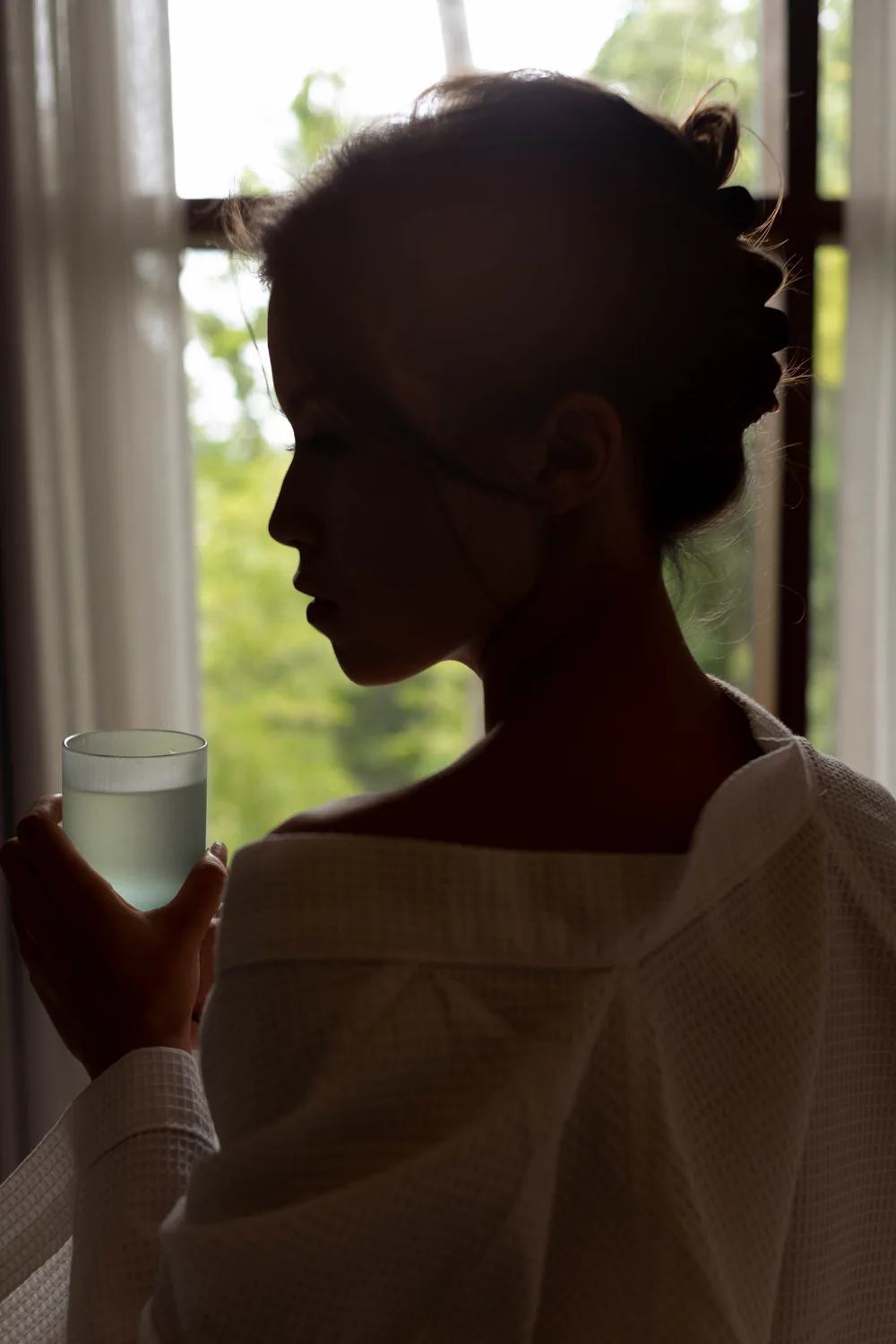A guest in a linen robe holds a glass of water by a window open to the trees